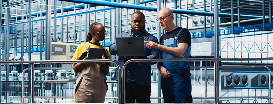 A consultant, engineer, and technician collaborate with a laptop on an industrial factory floor to define an MVP strategy.
