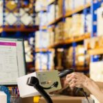 A warehouse worker in a yellow vest scans a package while sophisticated holographic data overlays visualize a new modern supply chain platform, featuring Docker containers, Kubernetes clusters, and 99.95% uptime enabled by Wishtree.